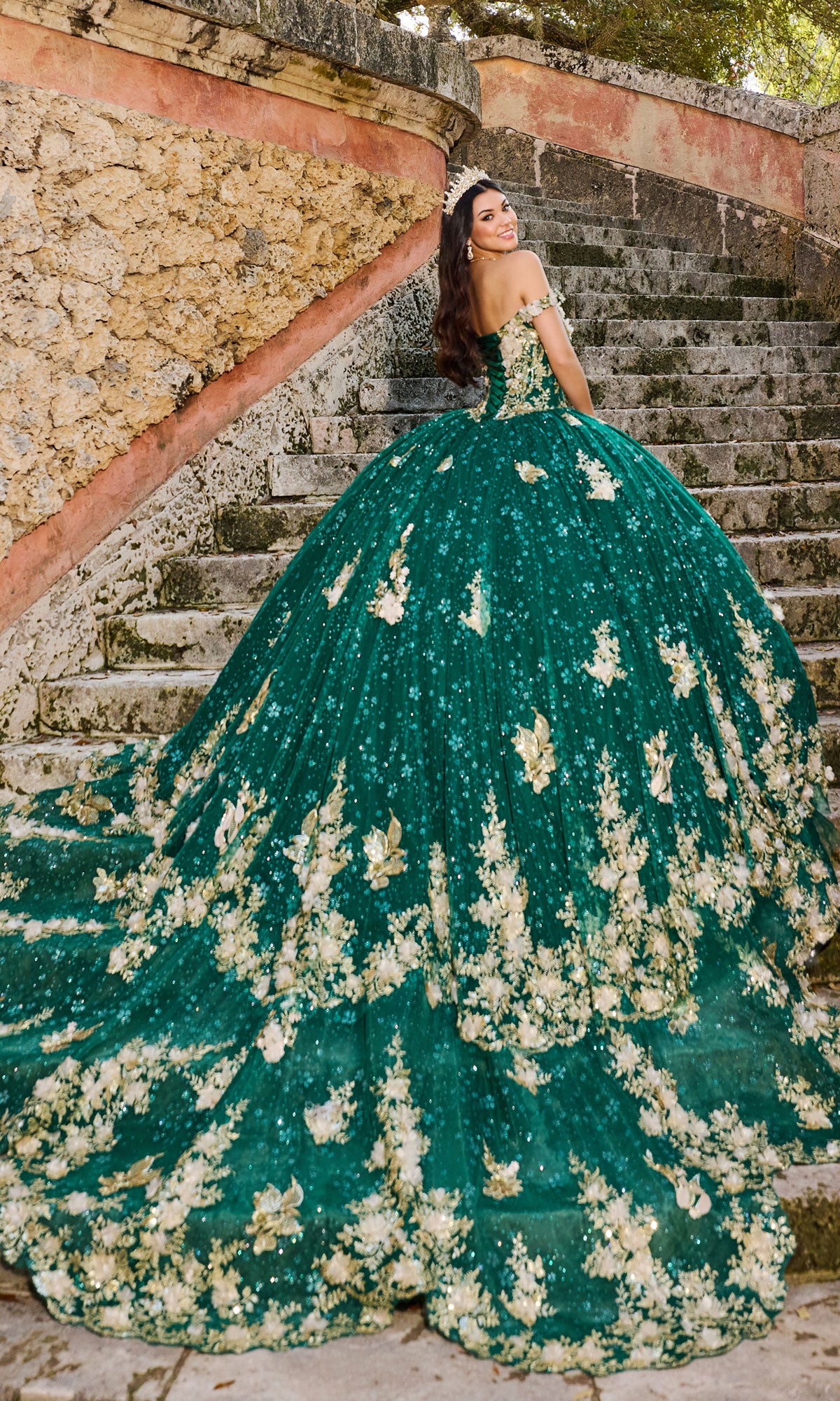 A woman in the Quinceanera Dress PR30111 by Princesa, a green gown with gold floral details and lace embroidery, stands on stone steps, smiling as she looks back over her shoulder.